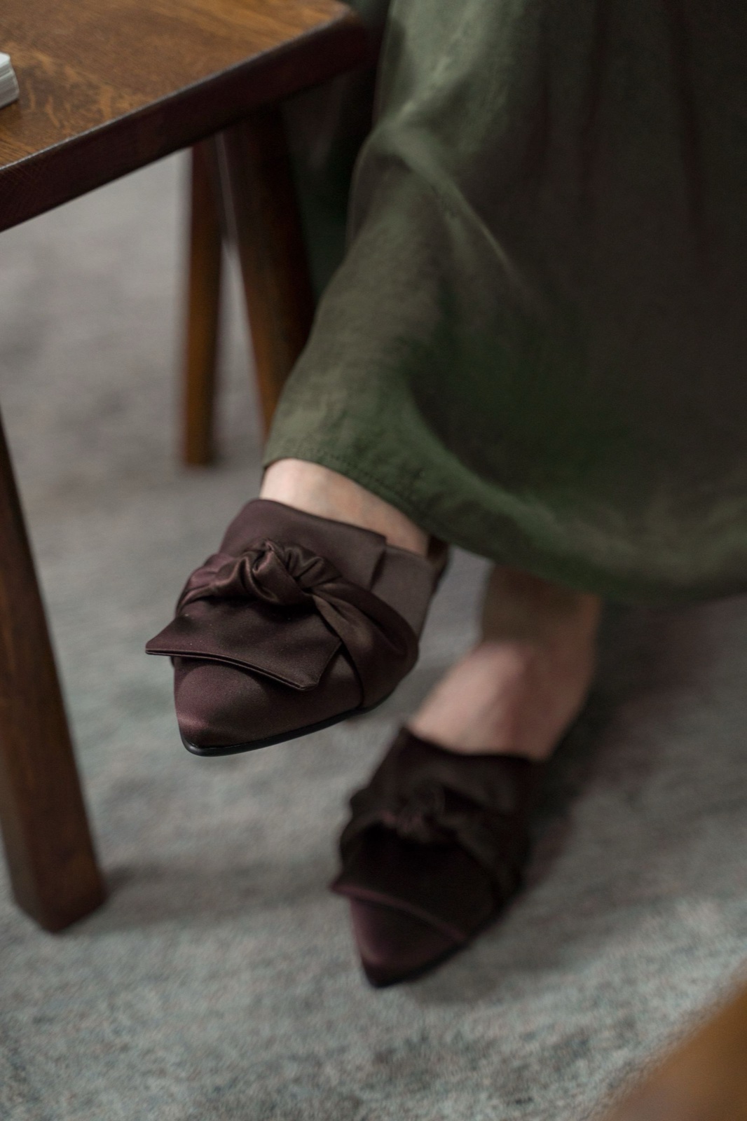 Pointed-toe brown satin mules with knotted bows worn with a long olive skirt, seen from above next to a wooden table