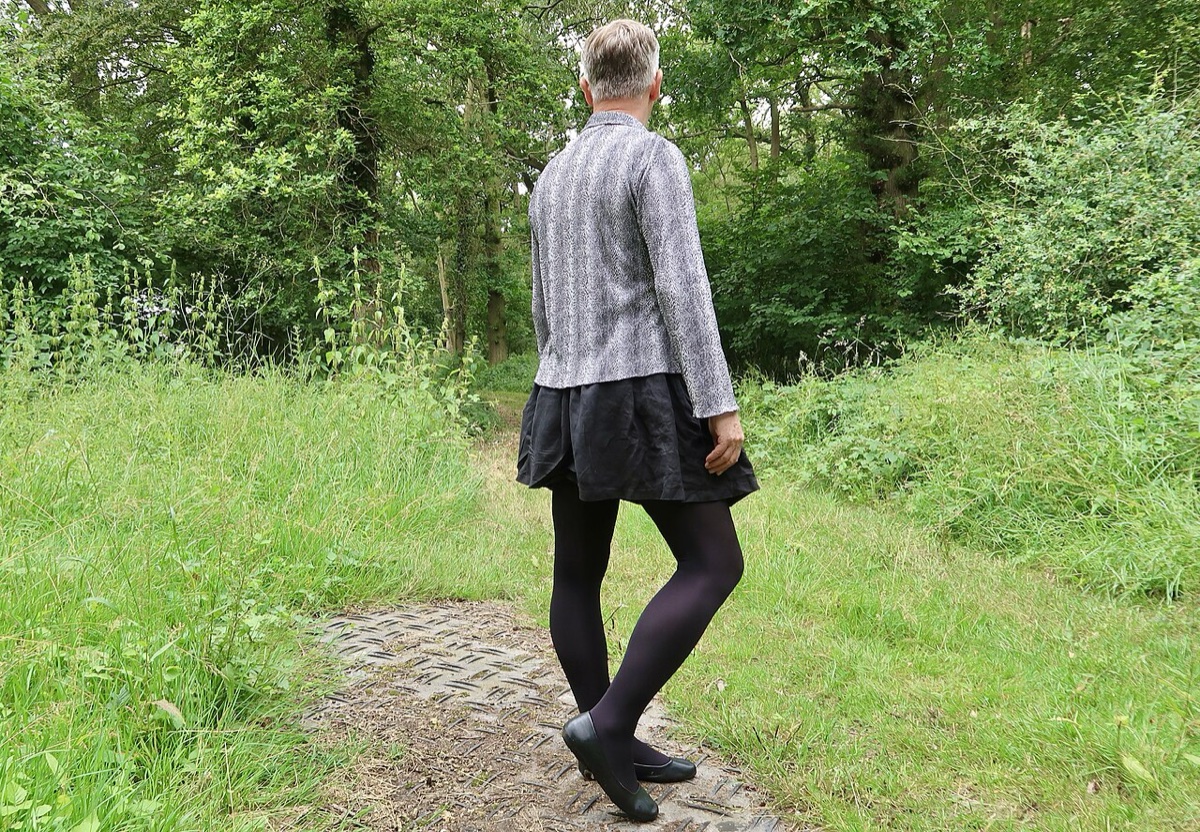 A person seen from behind walking a wooded path in a pleated skirt, black opaque tights, and plain flats