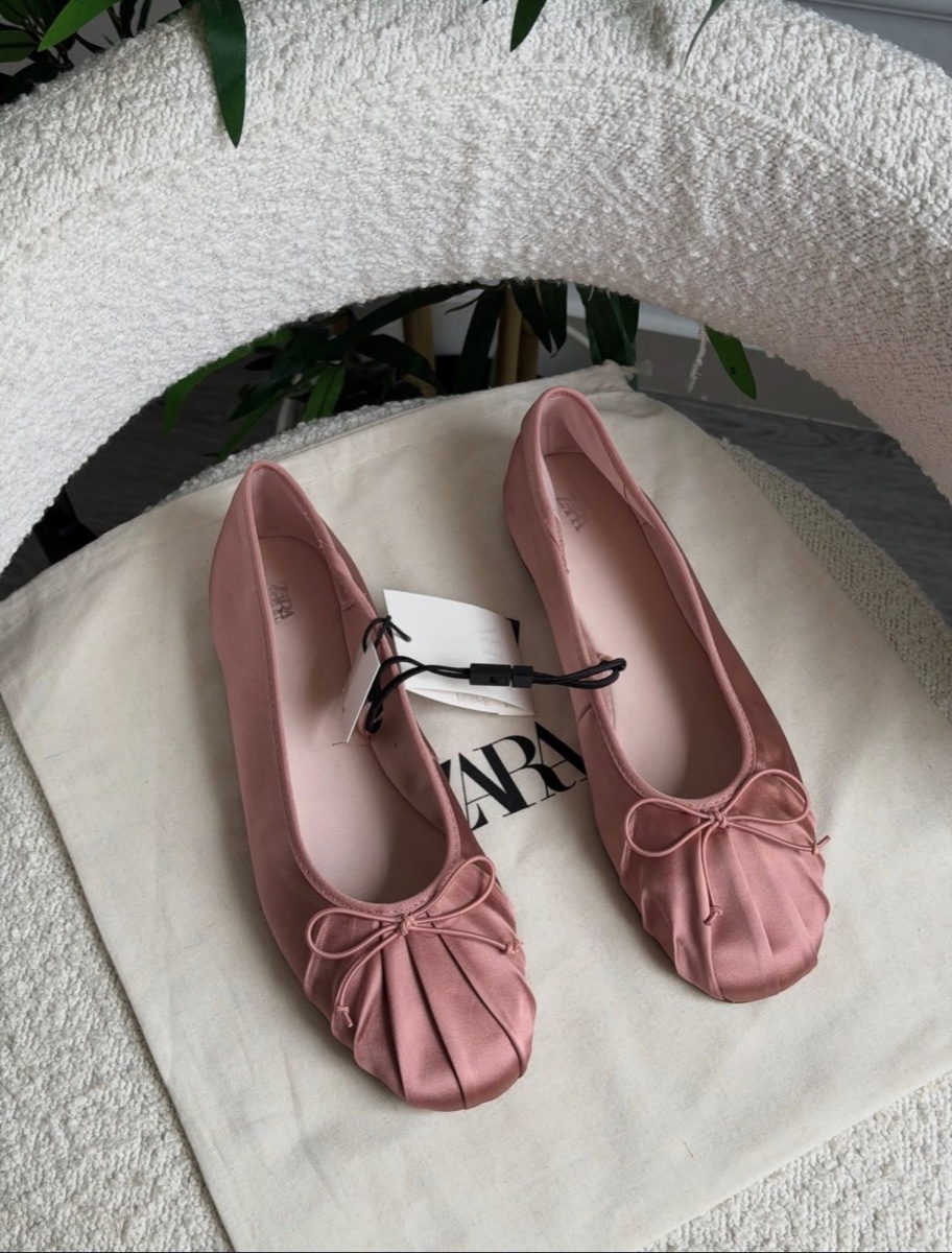 A pair of dusty pink satin ballet flats with bows and shirred toes, tags still attached, laid on a Zara dust bag on a boucle chair