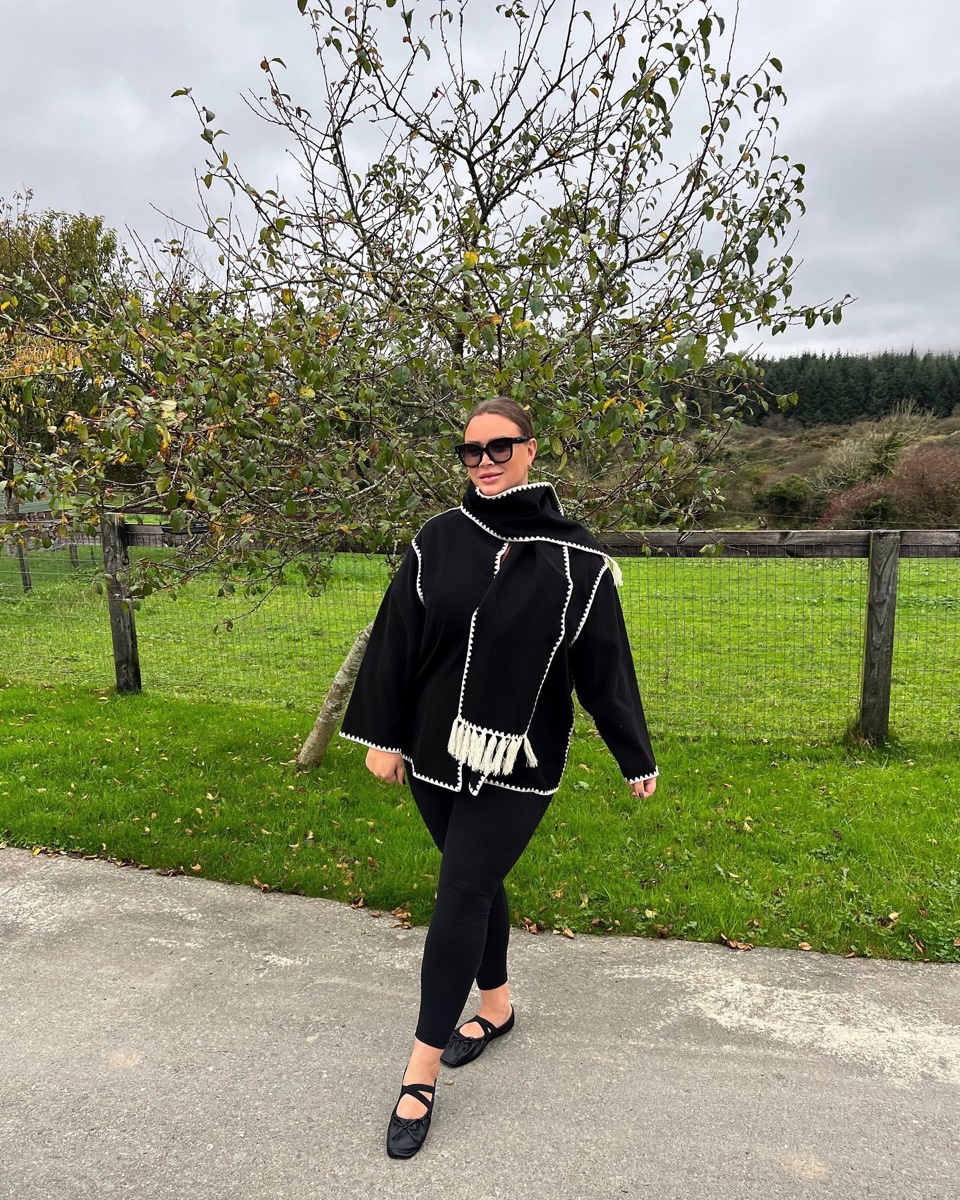 A woman in sunglasses walks on a path in a black scarf jacket with white whipstitch edges and tasseled attached scarf, black leggings and mesh flats