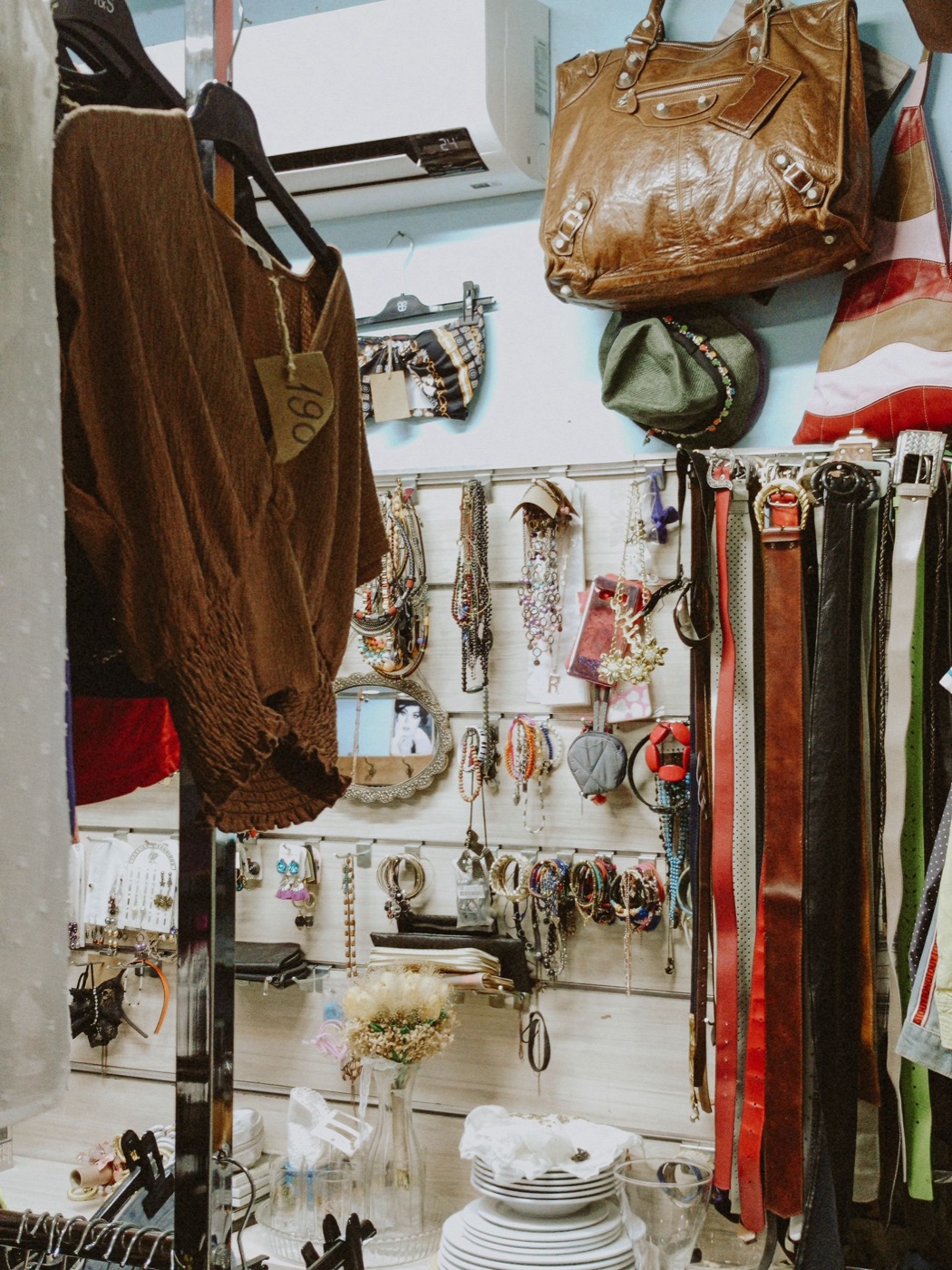 A brown crinkled top hanging in a secondhand shop with a handwritten cardboard price tag, beside a wall of belts and beaded necklaces