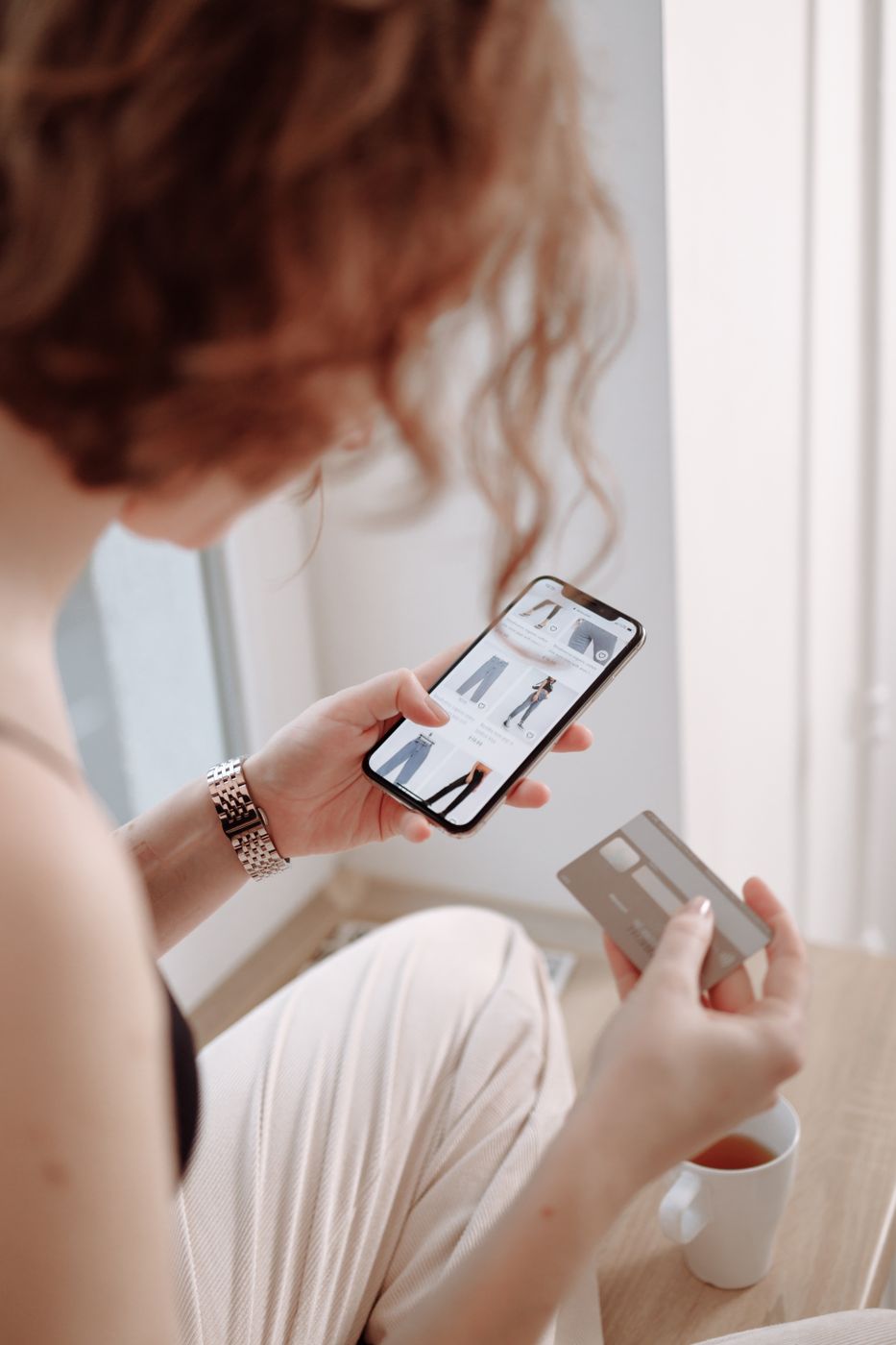 Woman holding a phone showing a shopping app grid in one hand and a credit card in the other