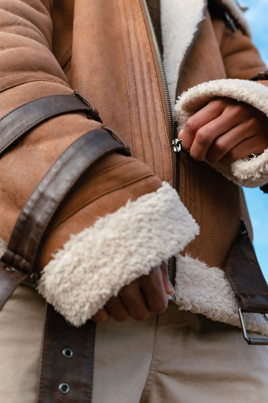 Hands zipping a tan suede jacket with thick cream pile showing at the cuff and along the opening