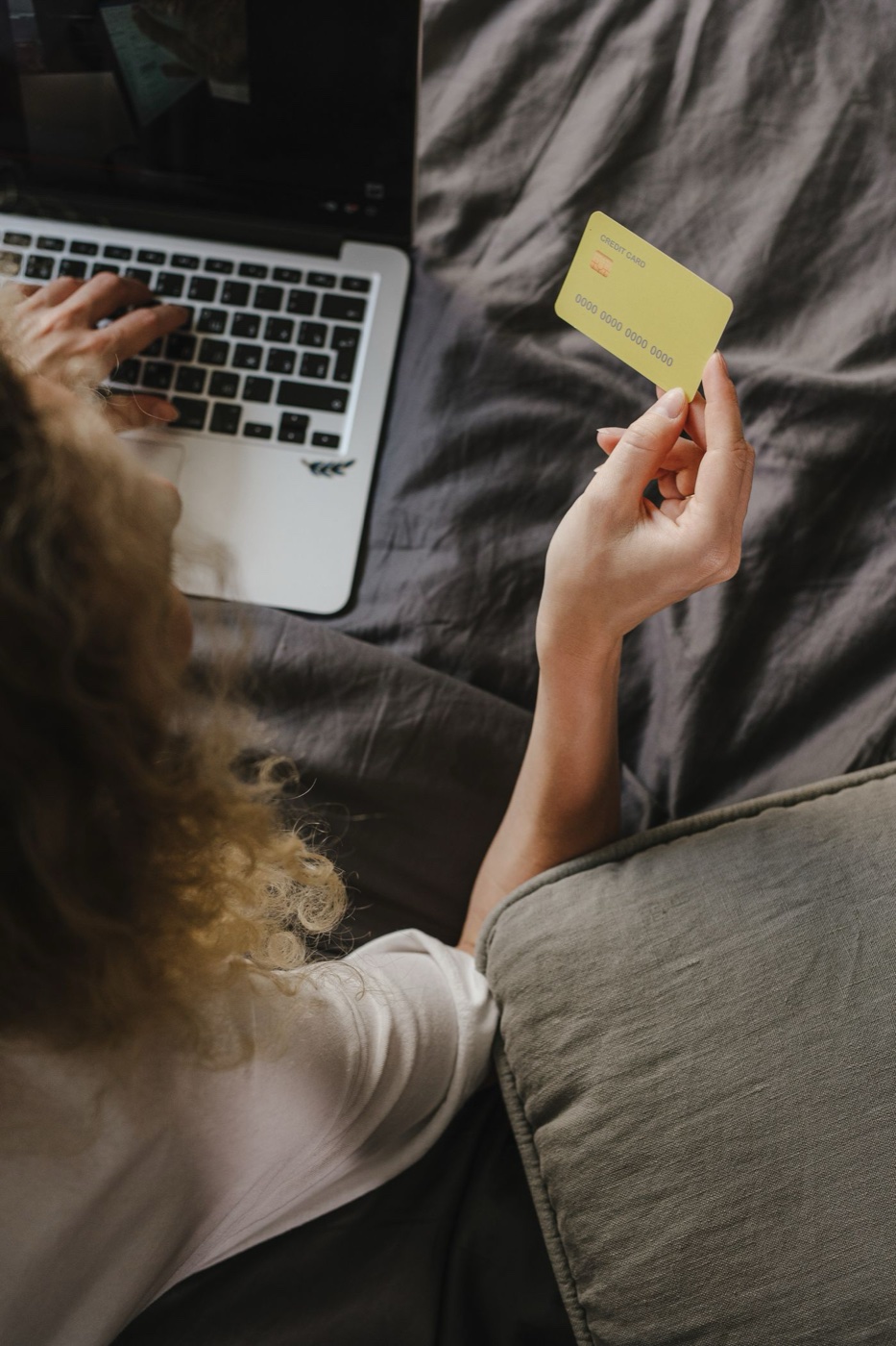 A shopper seen from behind holds a pale yellow credit card while typing on a laptop in bed