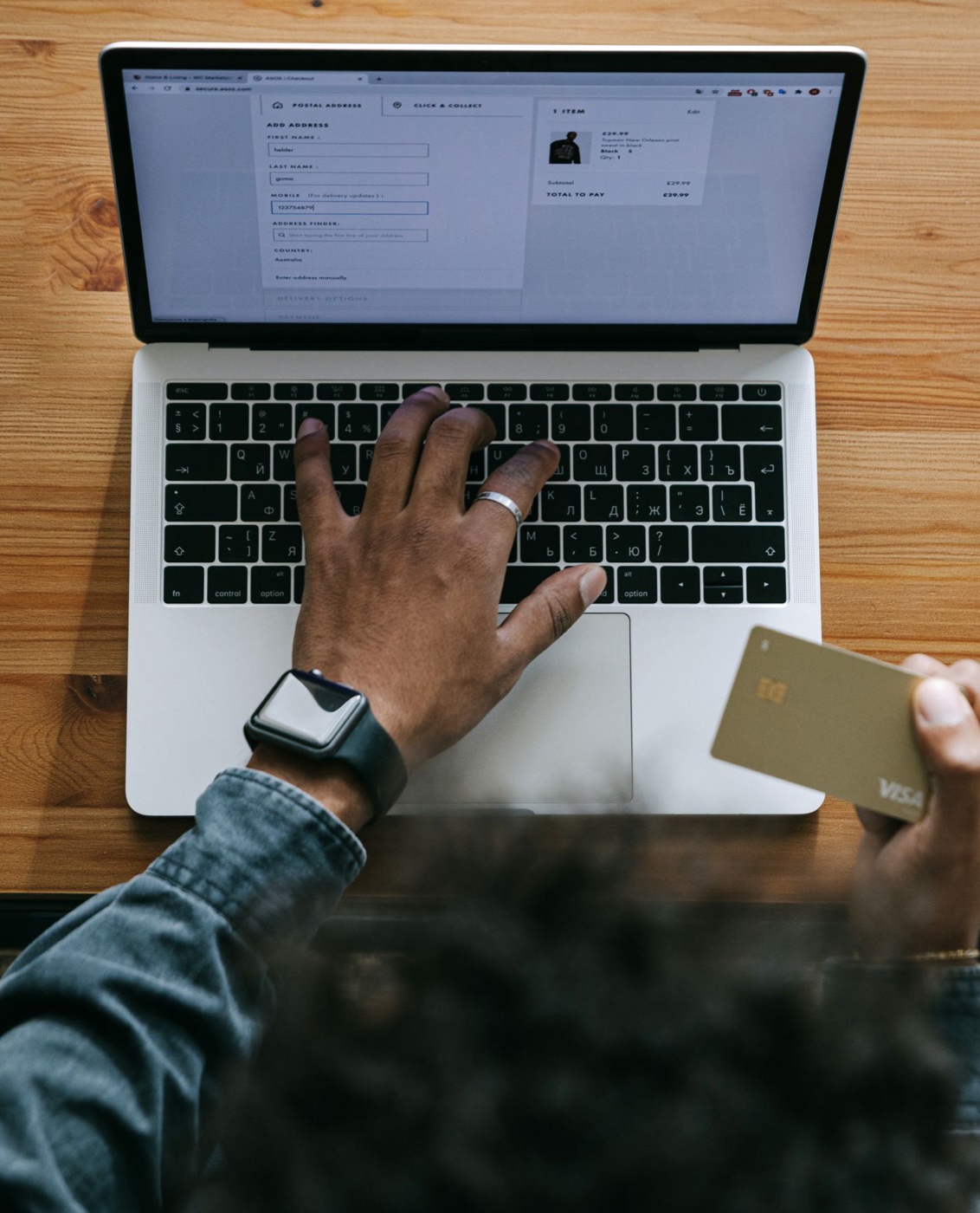 Overhead view of hands at a laptop showing a fashion checkout page, one hand holding a gold credit card