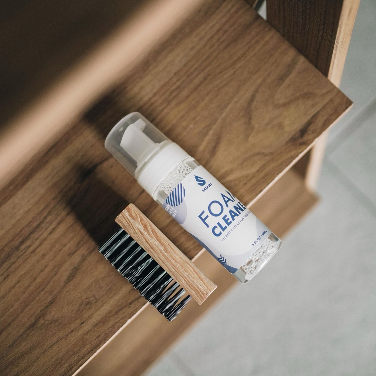 Bottle of foam shoe cleaner and a wooden bristle brush lying on a wooden shelf