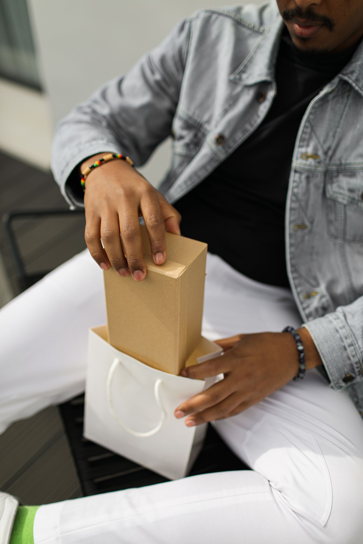 A man in a denim jacket pulling a kraft box out of a small white shopping bag on his lap