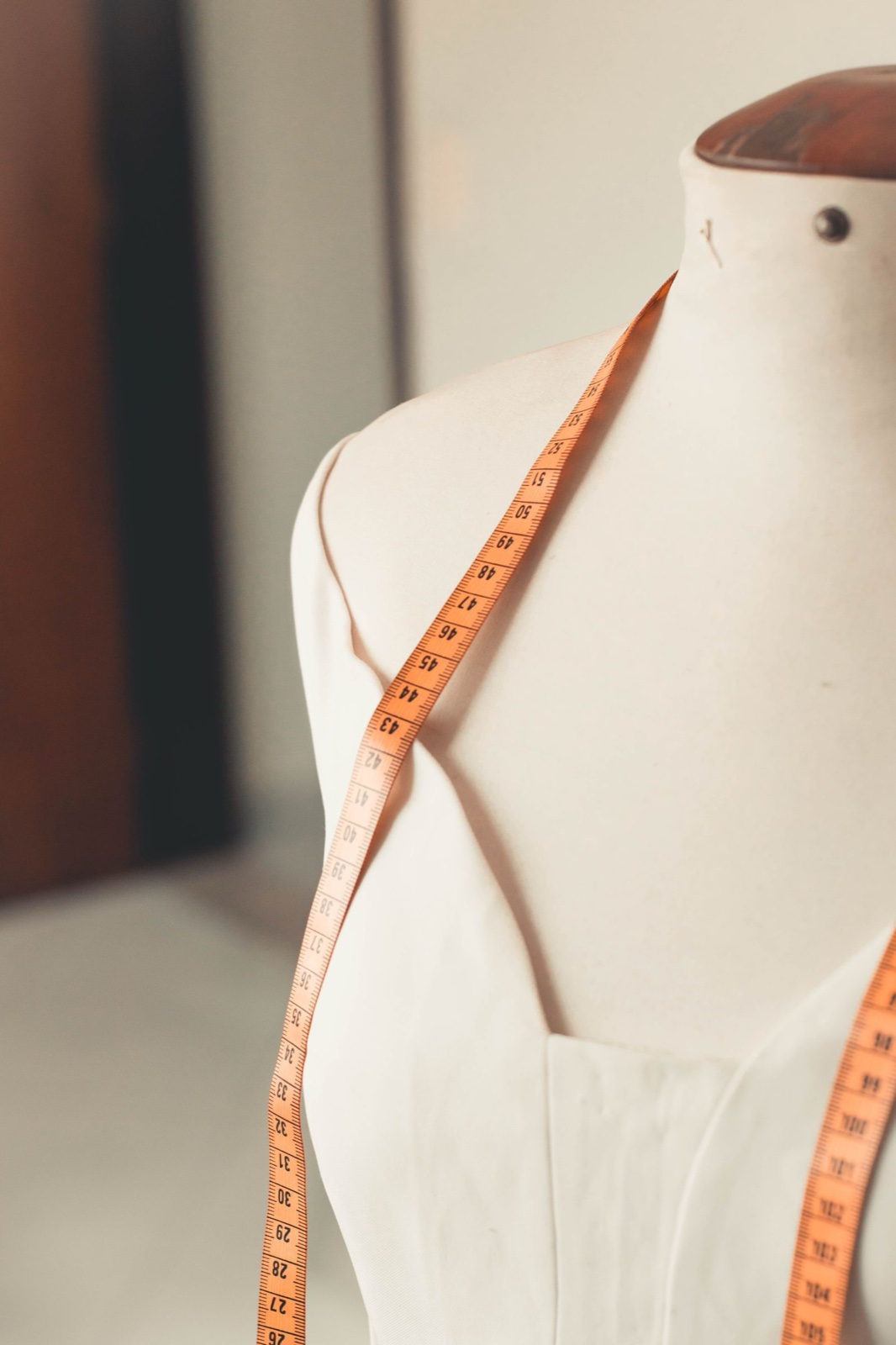 A dress form with an orange measuring tape draped over its shoulder, numbers facing out, in soft window light