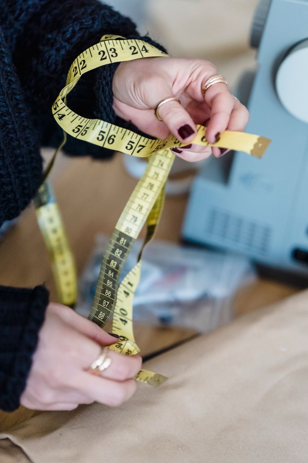 A tailor's hands holding a yellow measuring tape above a worktable with a sewing machine behind