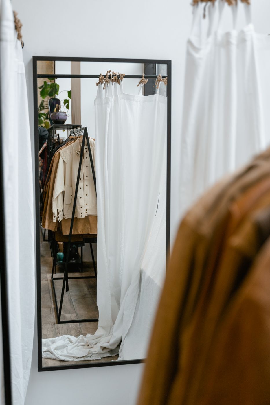 Full-length black-framed mirror on a white boutique wall reflecting an empty curtained fitting room and a rack of beige and camel jackets