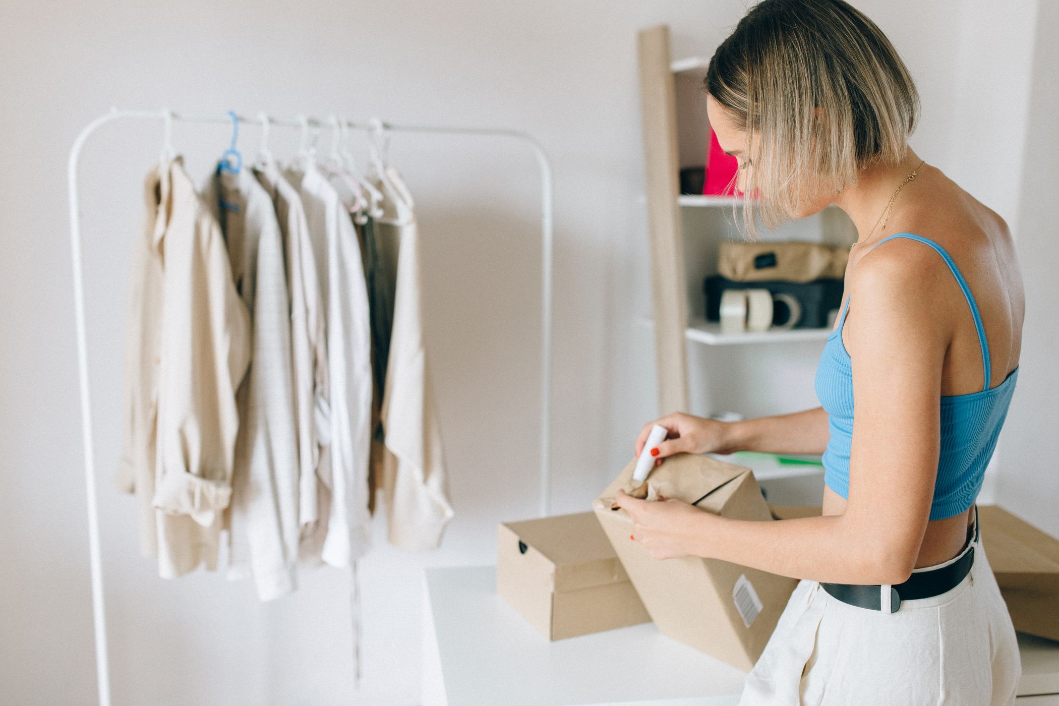 Woman in a blue top writing on a brown paper parcel over an open cardboard box, with a rail of pale shirts behind her