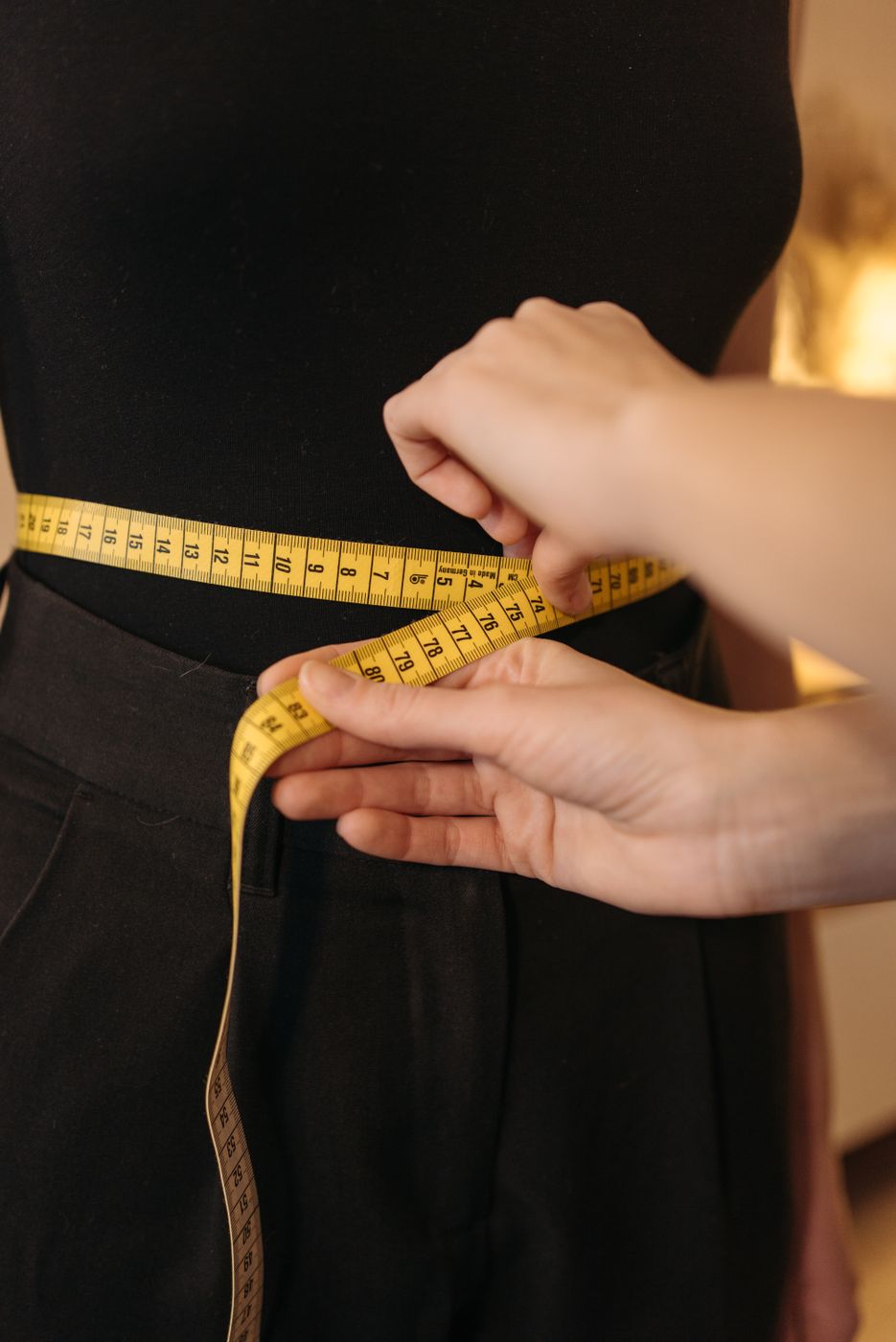 Close-up of two hands drawing a yellow tape measure around the waist of a person in black trousers and a black top