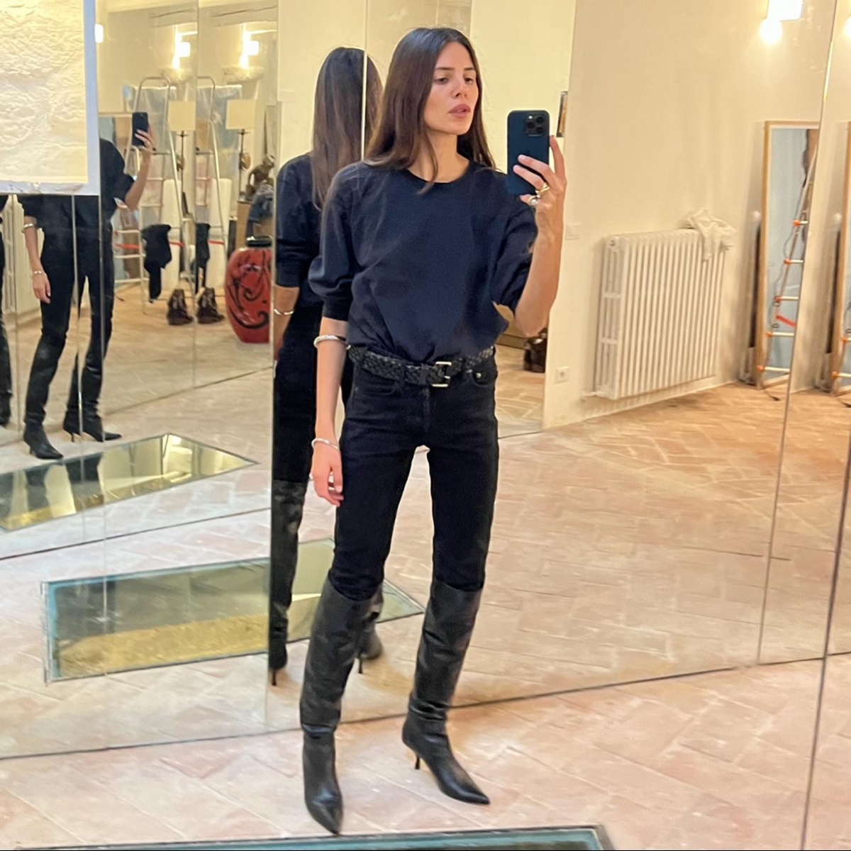 Mirror selfie of a woman in a navy sweatshirt, braided belt and black skinny jeans tucked into black knee-high boots, standing in a mirrored room