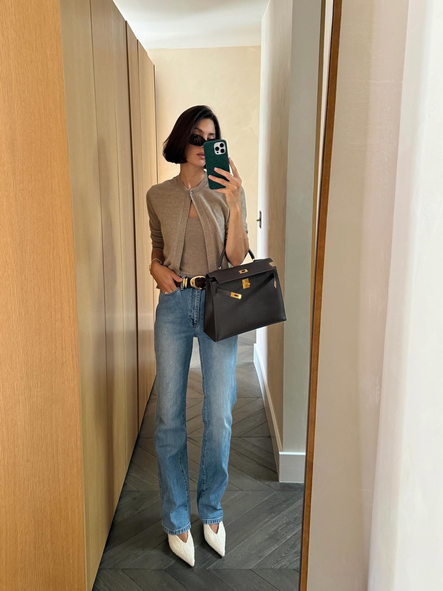 Mirror selfie in a wood-panelled hallway, woman in a taupe cardigan set and light-wash slim straight jeans holding a dark brown structured bag