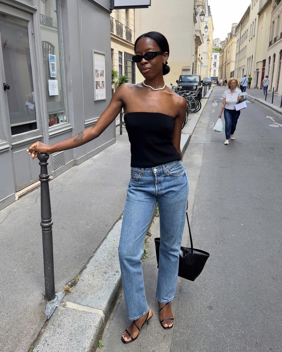 Woman in a black bandeau top, pearl necklace and mid-blue straight-leg jeans leaning on a bollard in a Paris street, black bag, strappy sandals