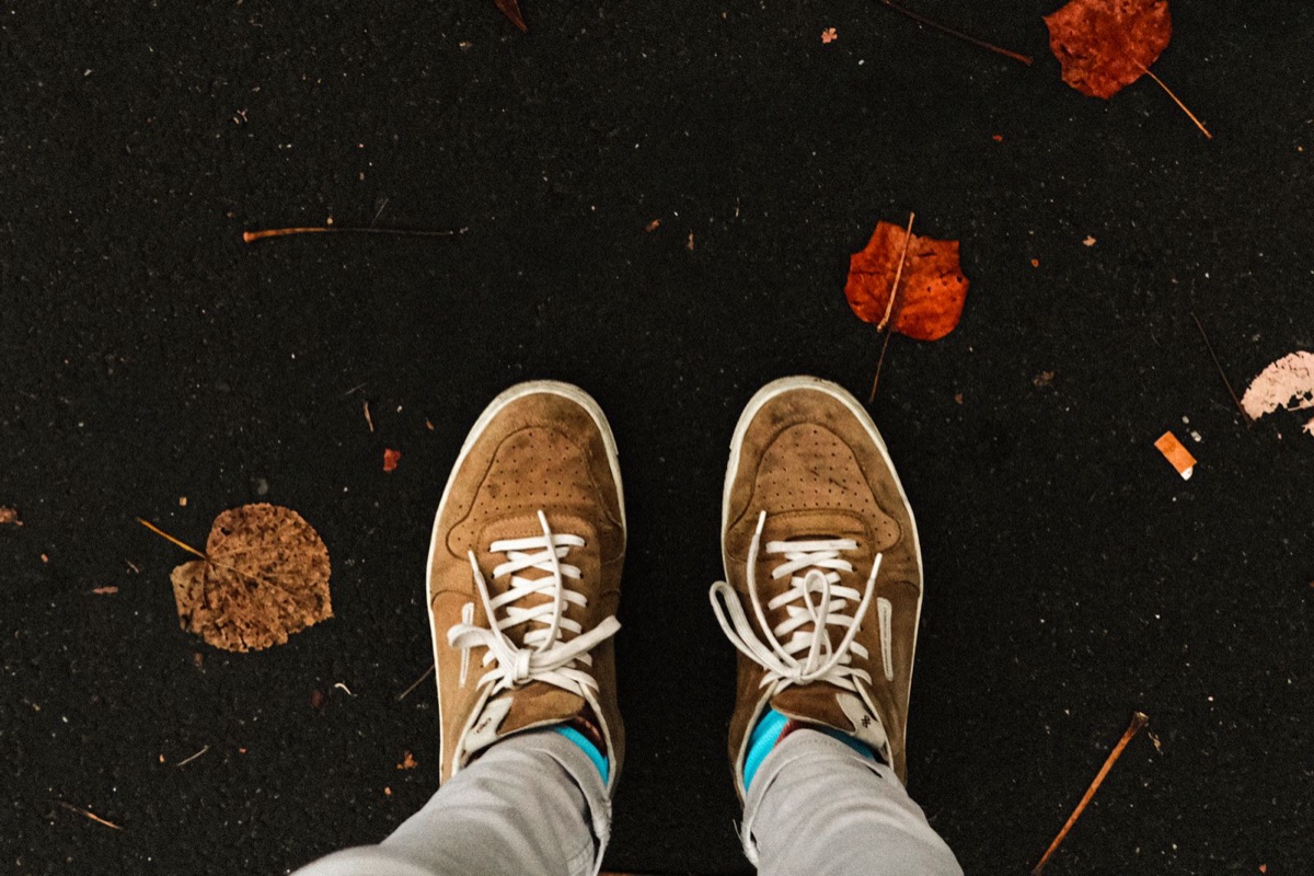 Overhead view of a person standing in tan suede sneakers on dark asphalt scattered with orange autumn leaves