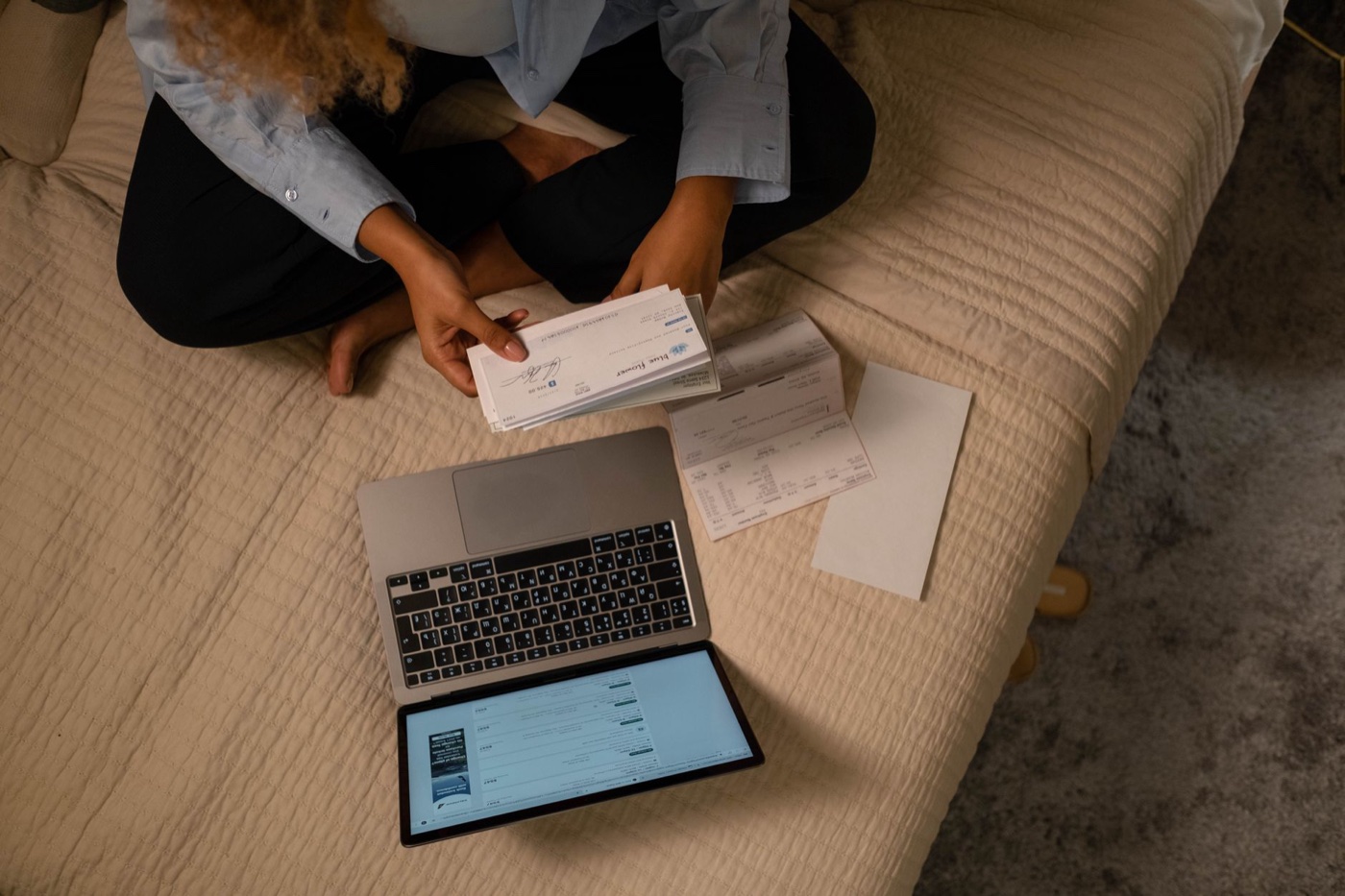 Overhead view of a woman on a bed reviewing paper checks and statements next to an open laptop
