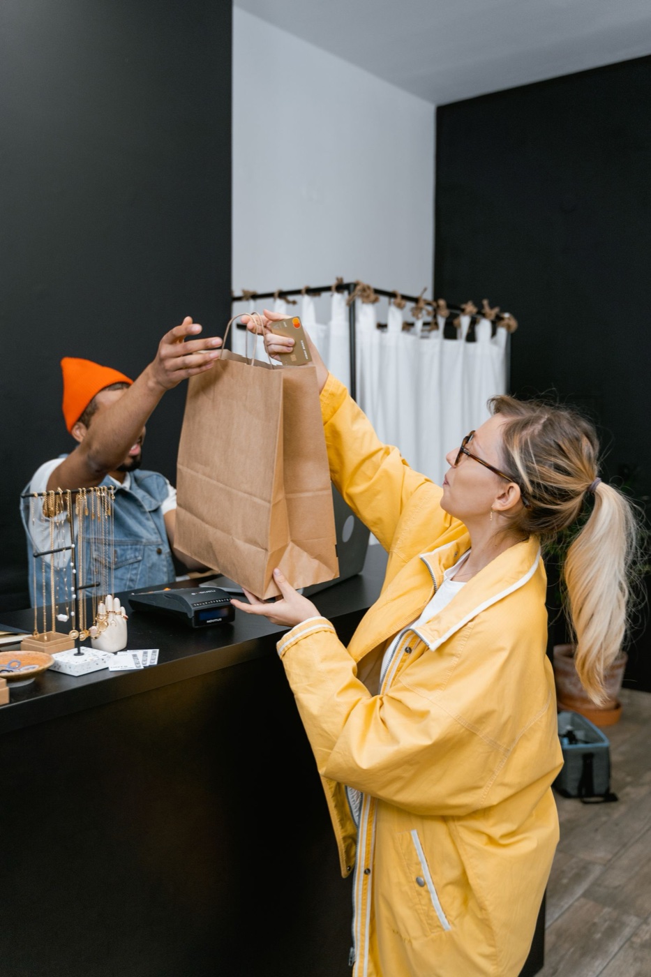 A customer in a yellow jacket taking a paper shopping bag across a dark counter from a sales assistant, a card still in her other hand