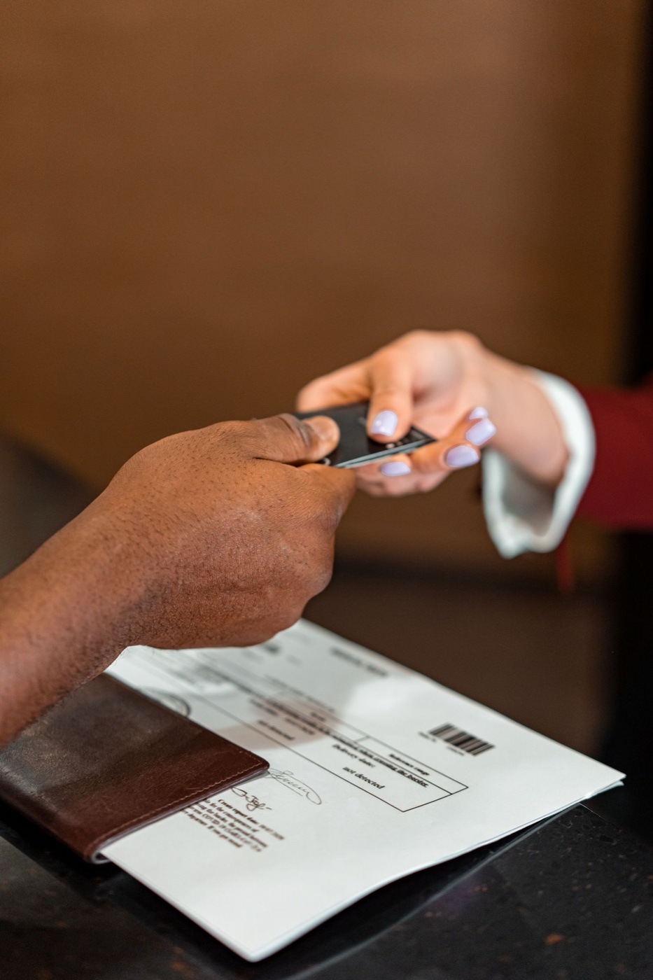 Two hands exchanging a dark payment card above a printed barcoded form and a leather folder on a dark counter