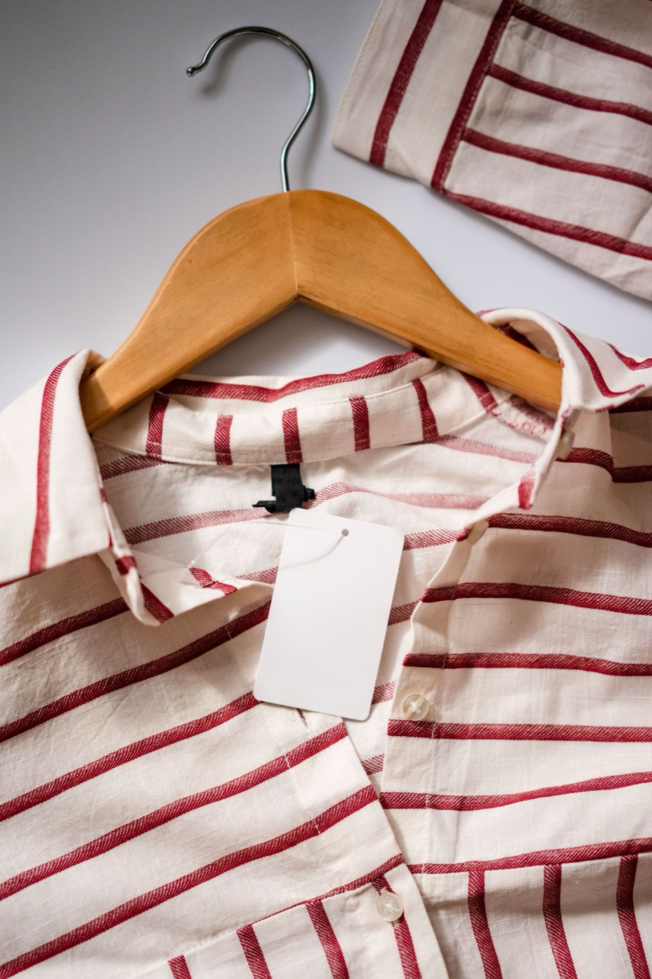A red and white striped cotton shirt on a wooden hanger with a blank white cardboard hangtag attached at the collar