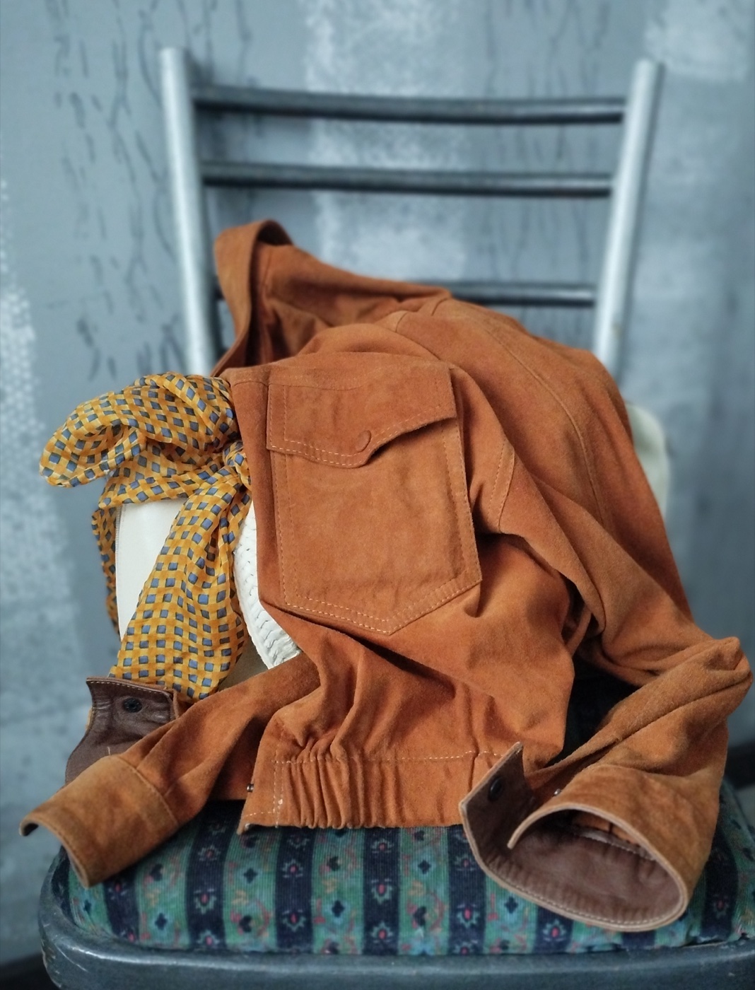 A tan suede jacket with a flap chest pocket and darker leather cuffs, draped over the back of a grey chair against a concrete wall