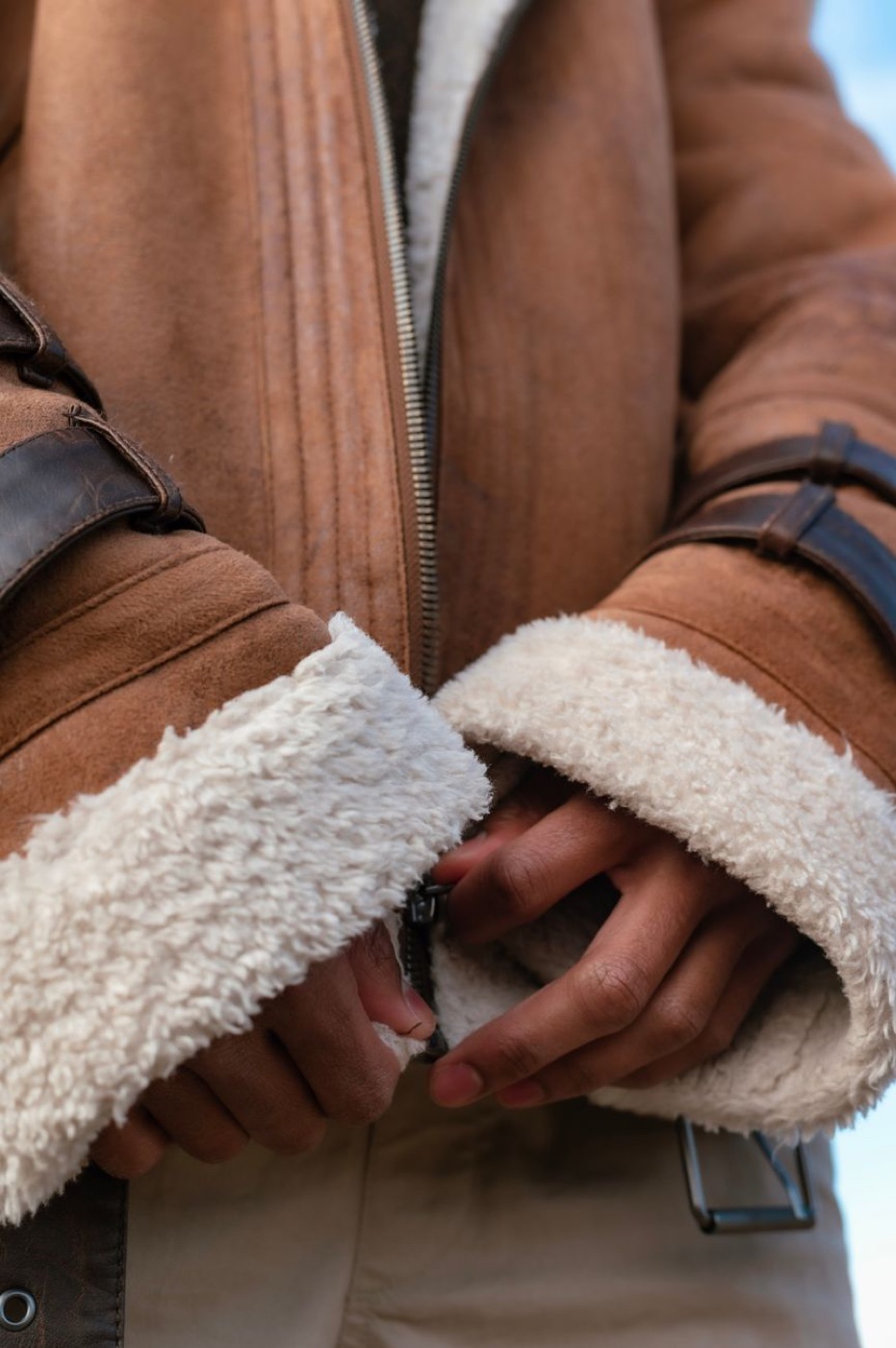 Close-up of hands doing up the zip on a tan suede jacket with thick cream shearling lining at the cuffs and collar