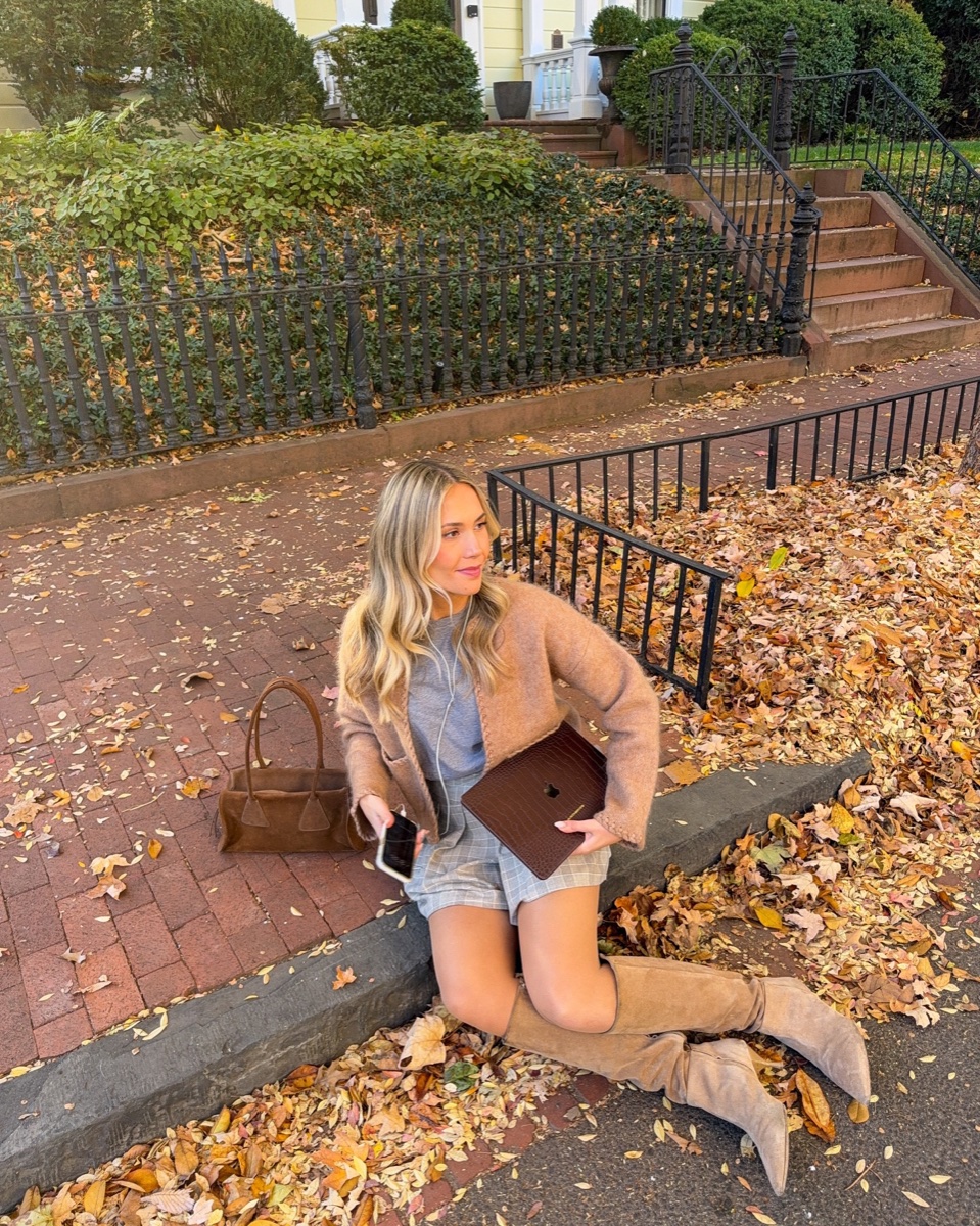 Tan suede knee-high boots stretched across a curb covered in fallen leaves, worn with a plaid mini skirt and camel cardigan on a brick sidewalk