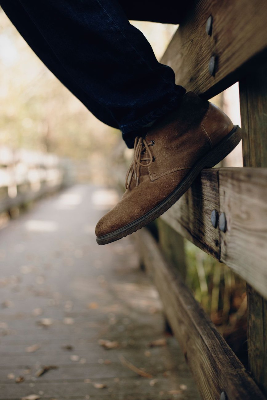 Close-up of a tan suede chukka boot resting against a wooden fence rail on an autumn boardwalk scattered with fallen leaves
