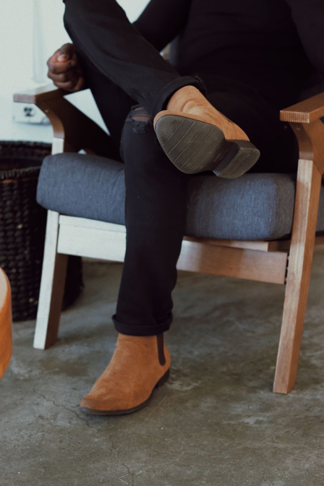 Person in black jeans sitting in a wooden armchair with one tan suede chelsea boot crossed over the knee, sole facing the camera