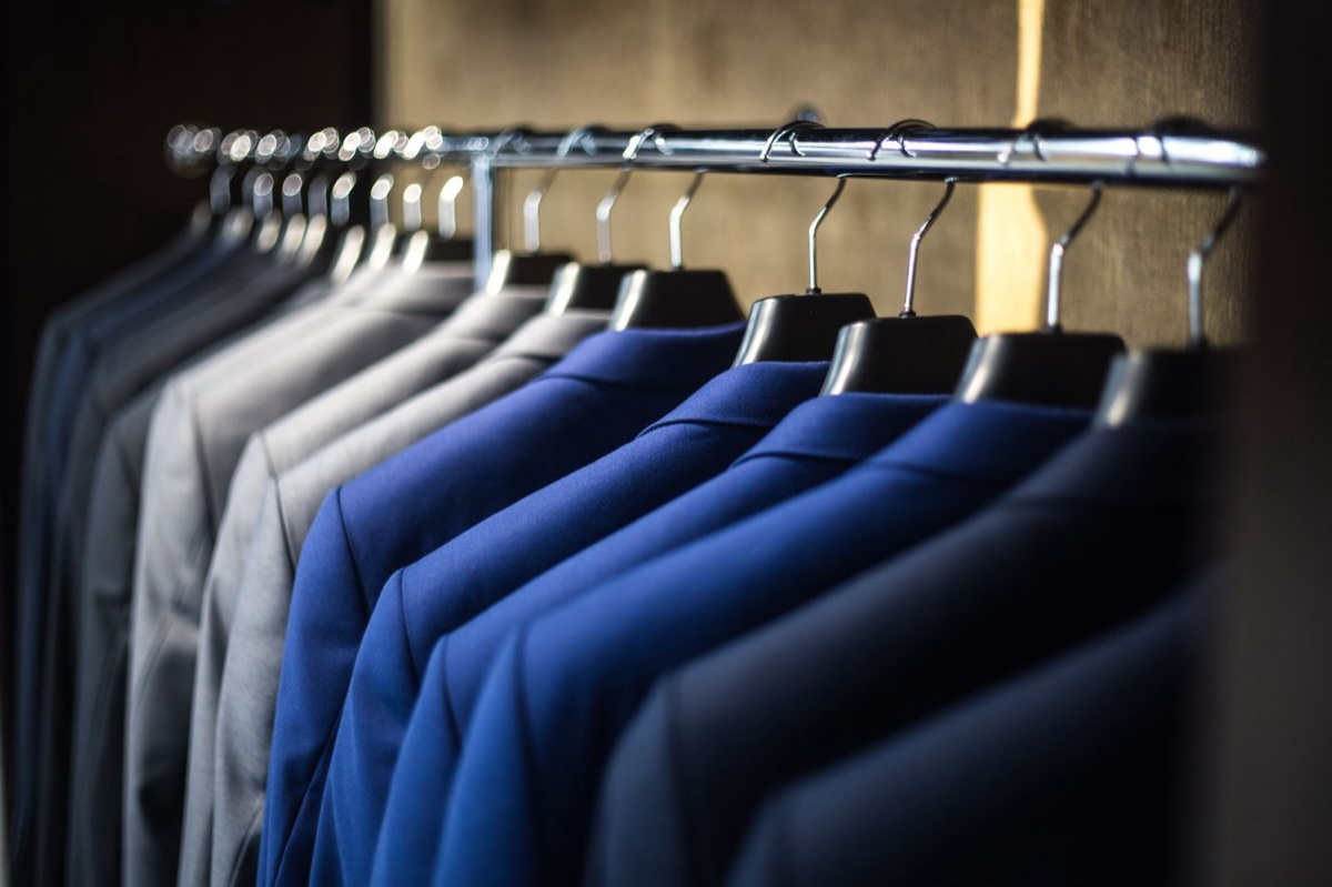 A close row of tailored suit jackets in grey, cobalt and navy hanging on wooden hangers along a metal rail