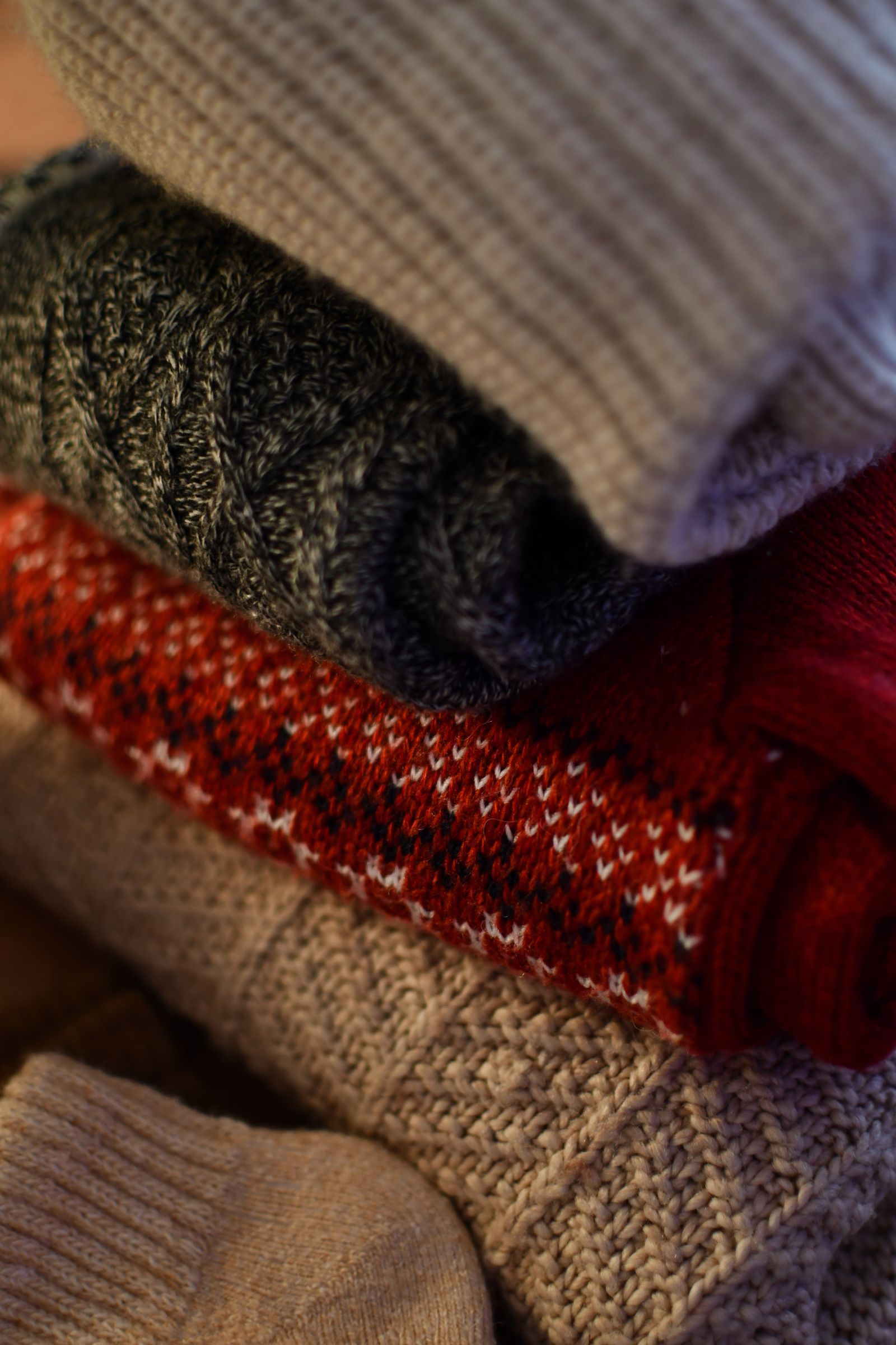 Stack of folded winter sweaters in warm colors, a ribbed oatmeal knit on top of charcoal cable knit and a red fair isle pattern