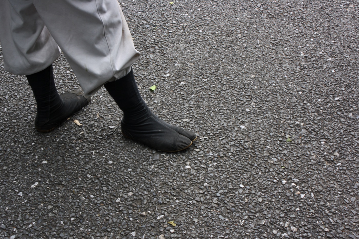 Person in worn black jika-tabi boots and white work trousers walking across wet asphalt