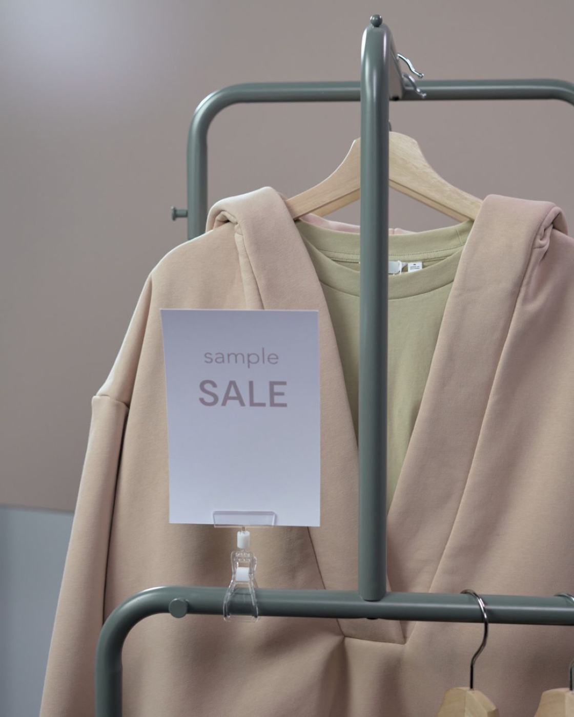 A beige jacket and a pale green shirt on a slim green clothes rail with a small white card clipped to the rail reading sample SALE