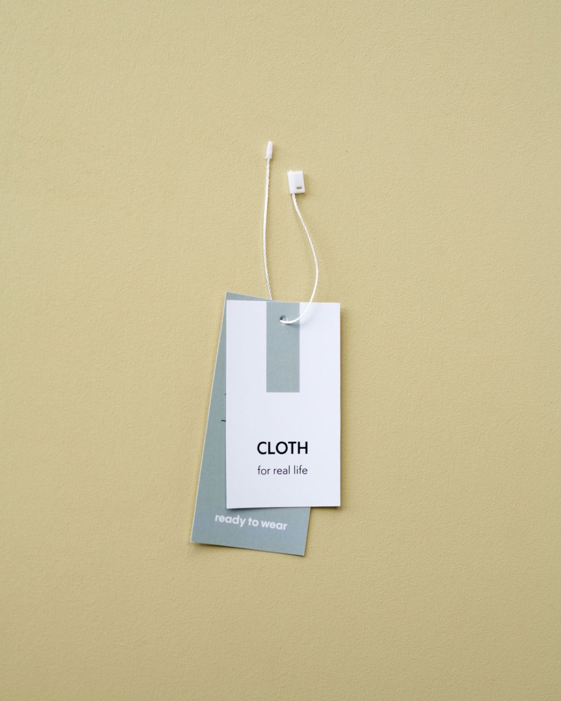 Two hang tags on a string against a mustard yellow wall, the front one printed with a brand name, the one behind it turned so its face cannot be read