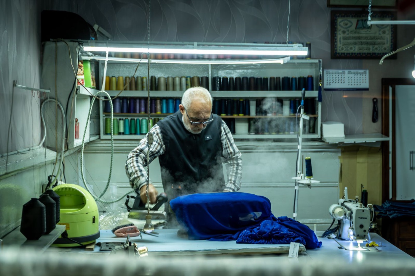 An older tailor pressing blue fabric with a steam iron in a workshop lined with spools of thread