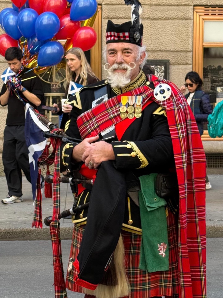 A piper in a red Royal Stewart kilt with a matching plaid pinned over the left shoulder, walking a New York street on Tartan Day