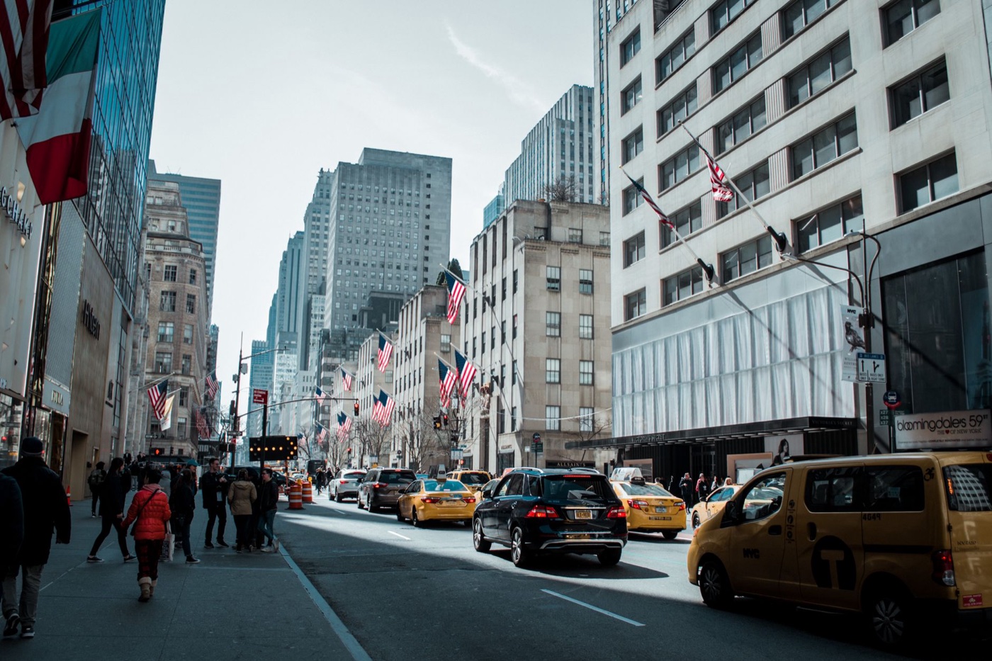 Fifth Avenue in New York with storefronts, American flags, pedestrians and yellow taxis