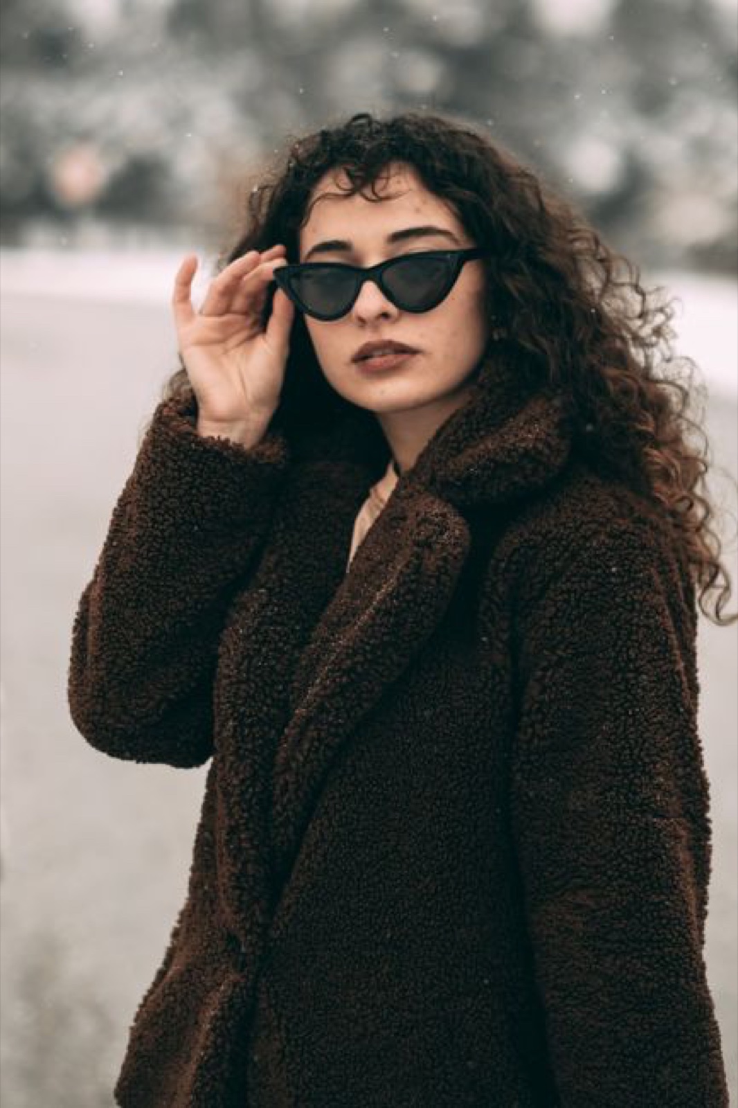 A woman in a dark brown bouclé teddy coat adjusting cat-eye sunglasses on a snowy road