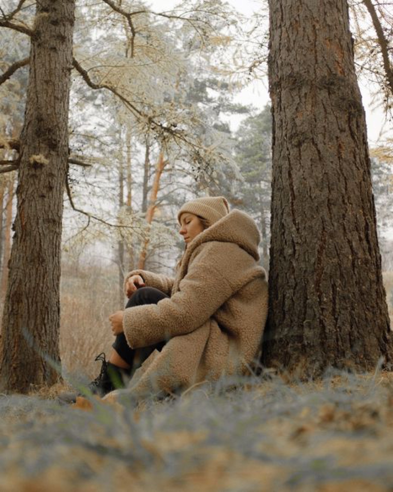 A woman in a camel teddy coat and matching beanie sitting against a pine trunk in a frosted autumn forest, face turned down in profile