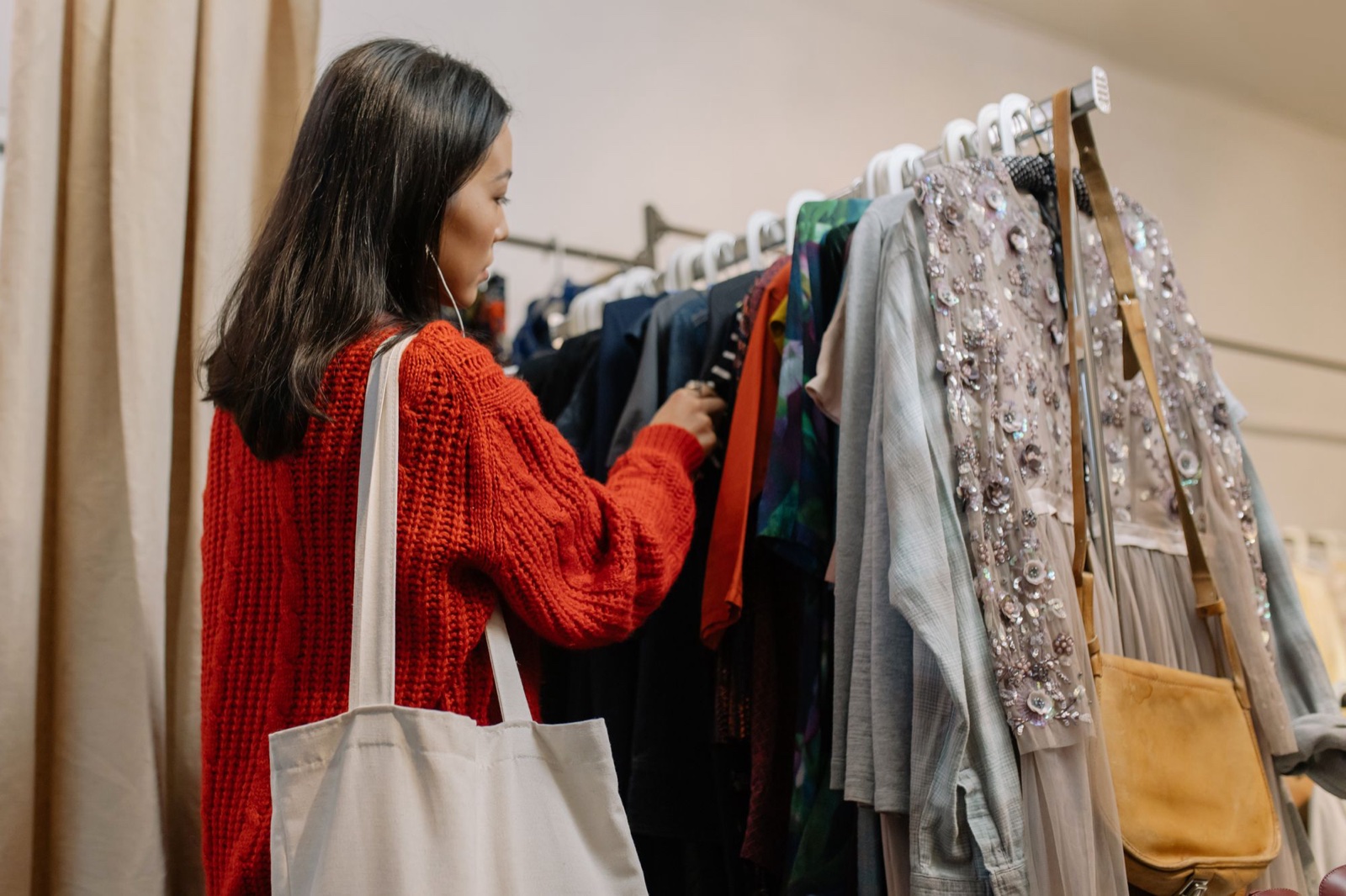 Woman in a red cable knit sweater with a canvas tote browsing a mixed rack of clothes in a secondhand store