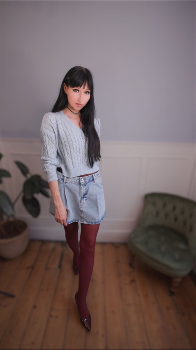 Person standing in a pale blue cable-knit sweater, pleated denim mini skirt, burgundy opaque tights and burgundy patent heels on a wooden floor