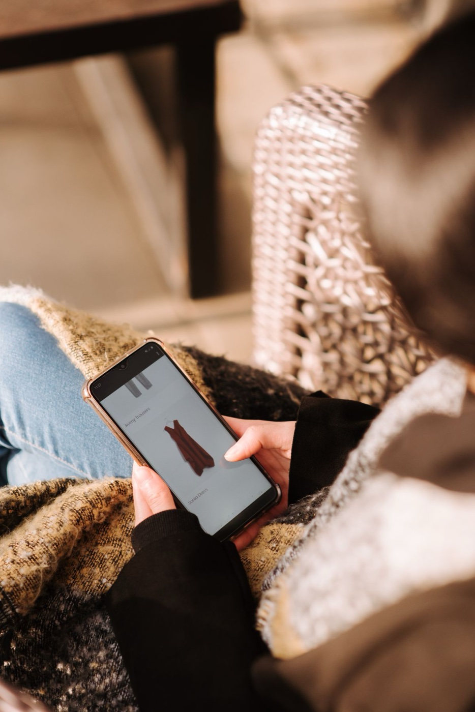 A person wrapped in a blanket browsing a clothing shop app on a phone, a dress listing on screen
