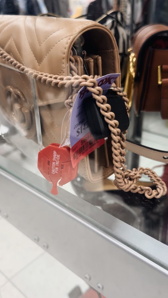 A beige quilted Gucci chain bag in a T.J. Maxx display case, purple Compare At tag and red $999 price sticker hanging from the strap
