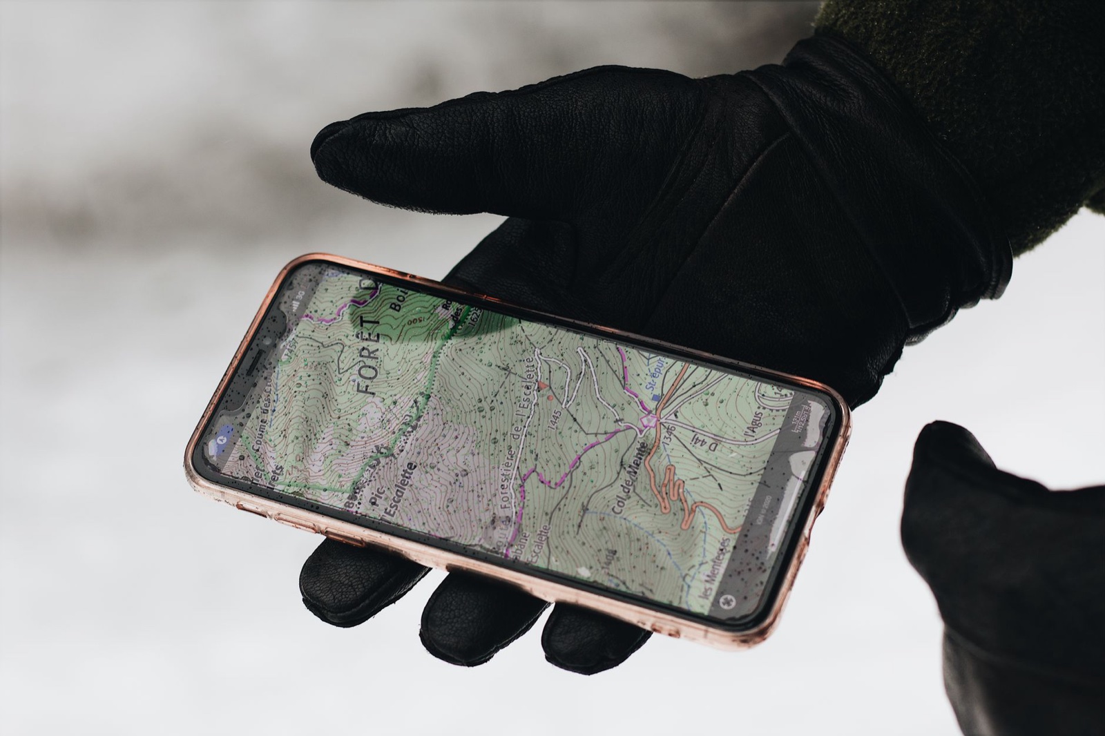 A black leather gloved hand holding a smartphone showing a topographic hiking map over a snowy background