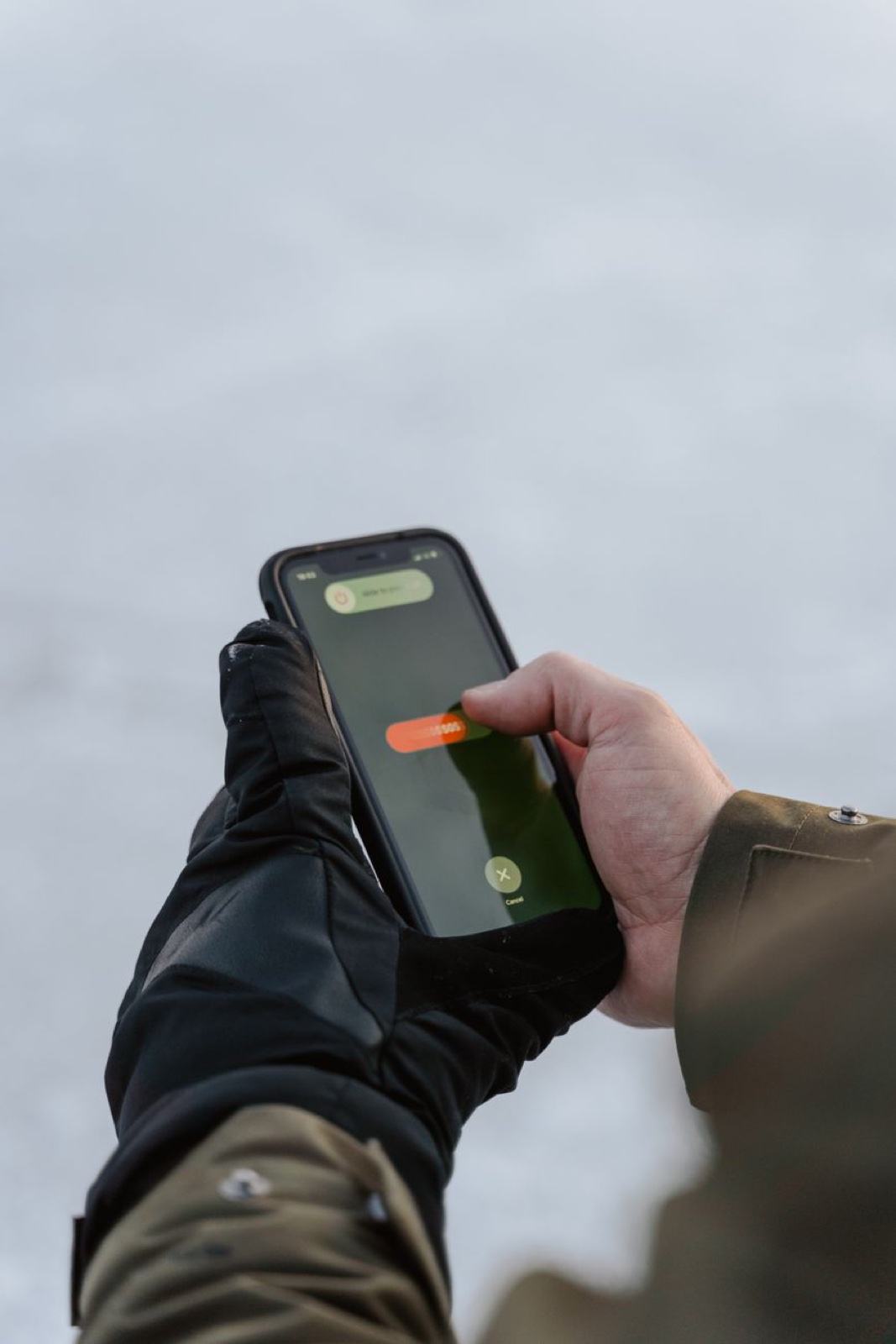 A hand pulled halfway out of a black insulated glove to press a smartphone screen outdoors in winter, the empty glove fingers folded back over the wrist
