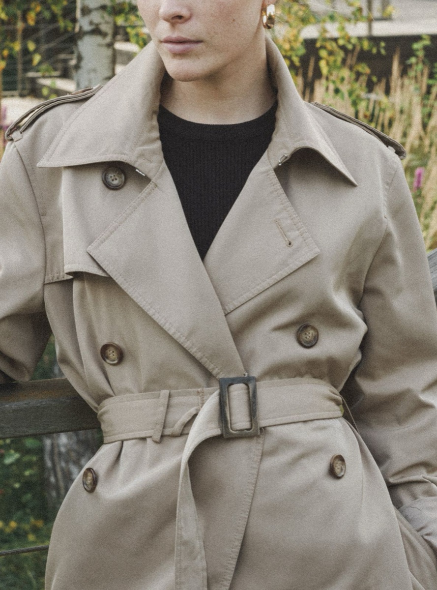 Torso of a belted beige double-breasted trench coat with tortoiseshell buttons and a horn buckle, photographed outdoors against autumn foliage