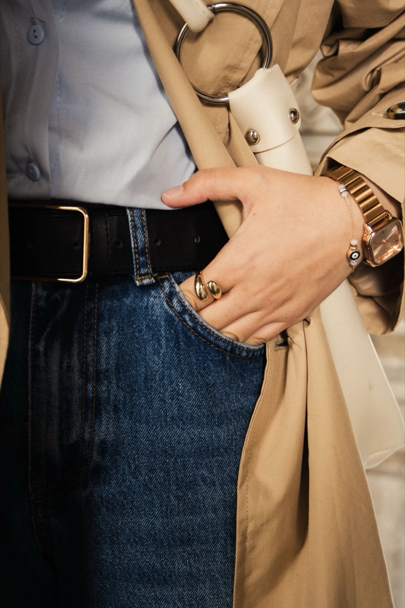 Close-up of a hand tucked into a jean pocket under an open beige trench coat, black belt with gold buckle, gold watch
