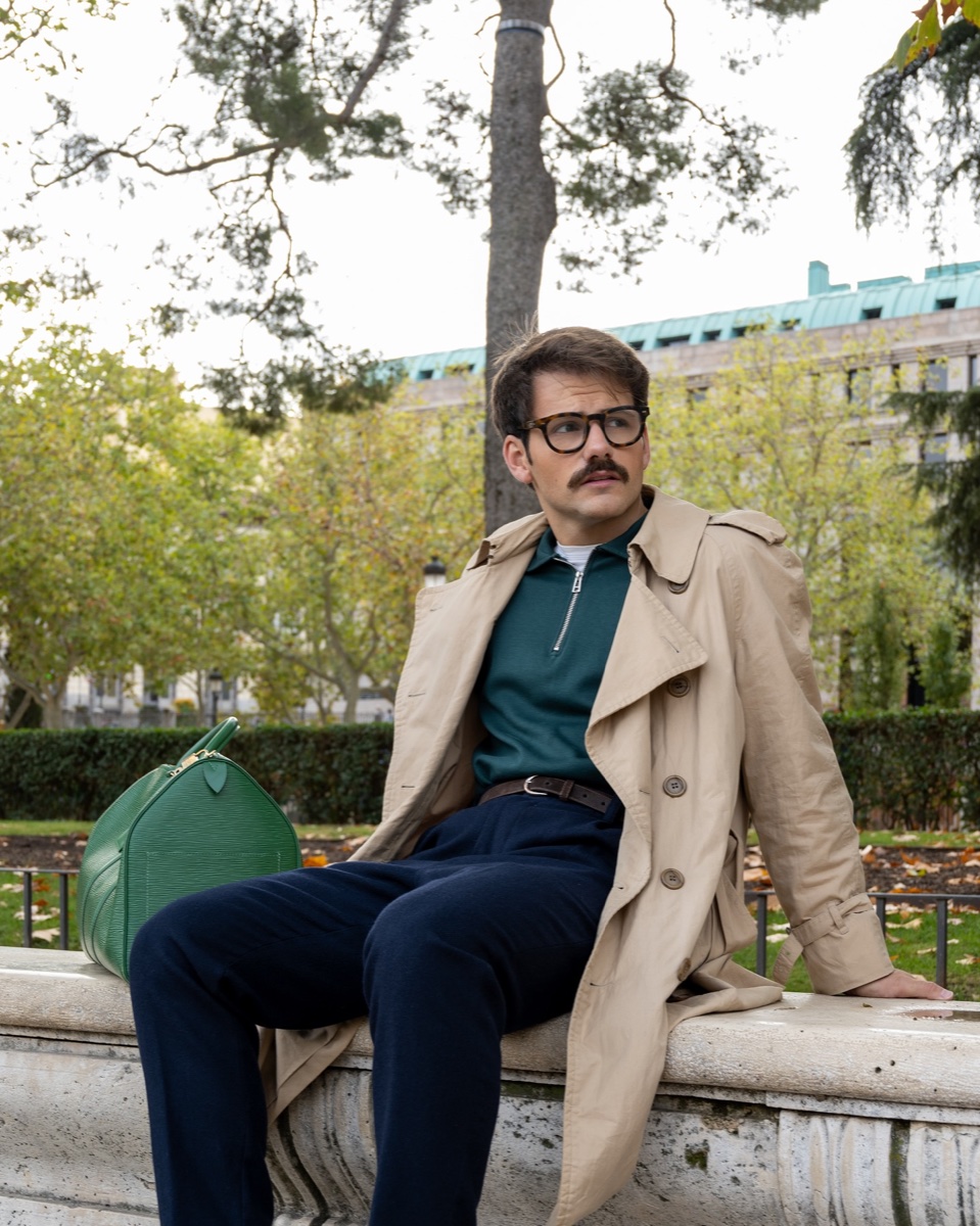 Man in glasses and a green zip polo sitting on a stone park wall in an old beige double-breasted trench, green bag beside him, autumn trees behind