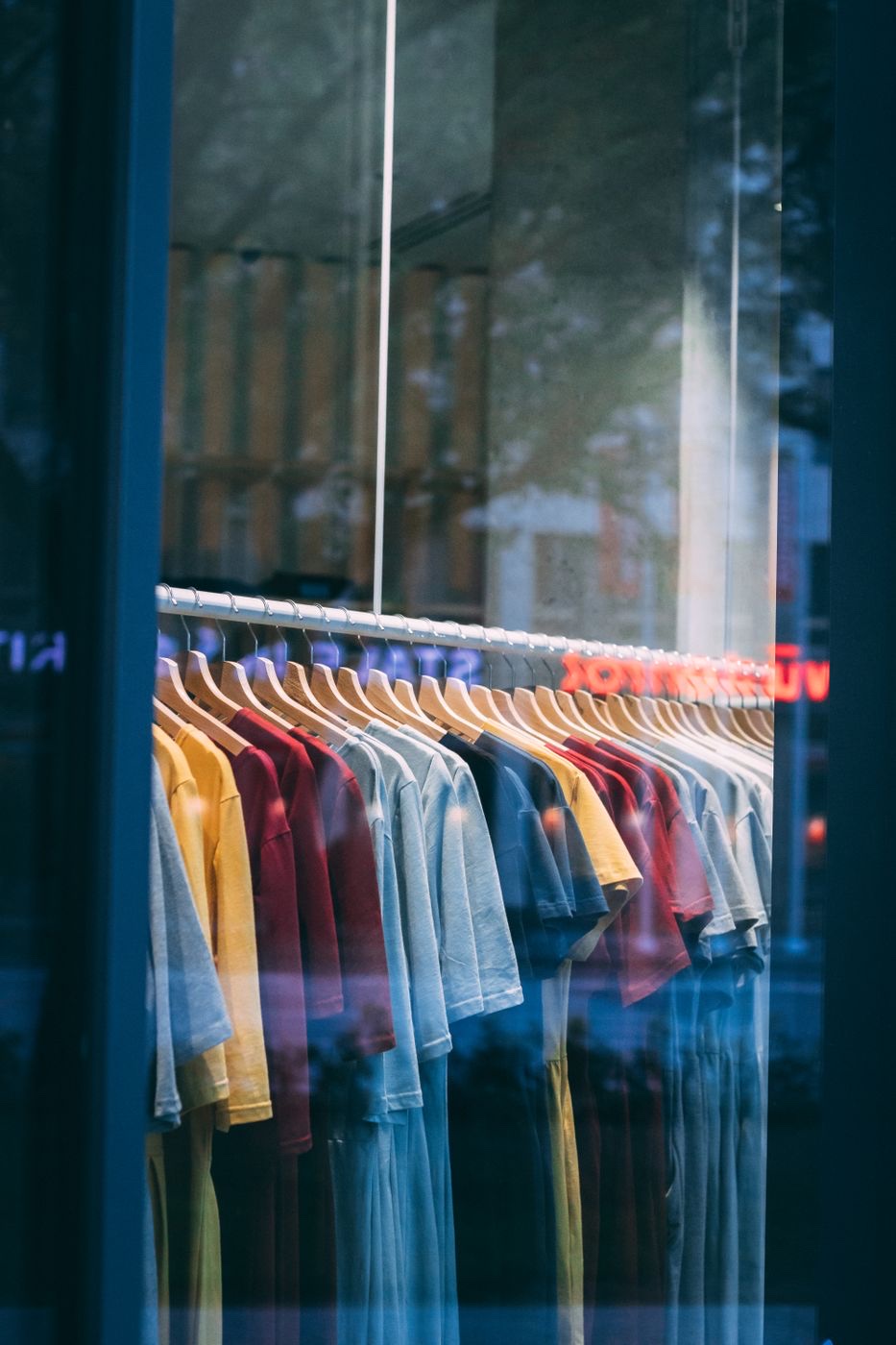 A rack of tee shirts in yellow, red, and blue on wooden hangers seen through a shop window at dusk, with street reflections across the glass