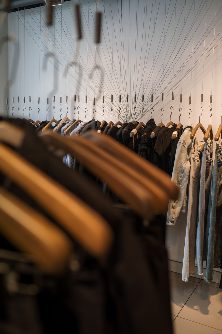 Two clothing rails in one frame, a blurred foreground rail of dark garments on wooden hangers and a sharp back wall rail holding denim